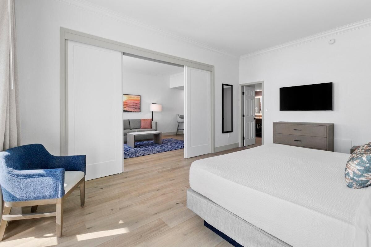 Camera d'albergo con un letto grande, una scrivania, una sedia, una TV e vista su un'altra stanza con un divano e un quadro.