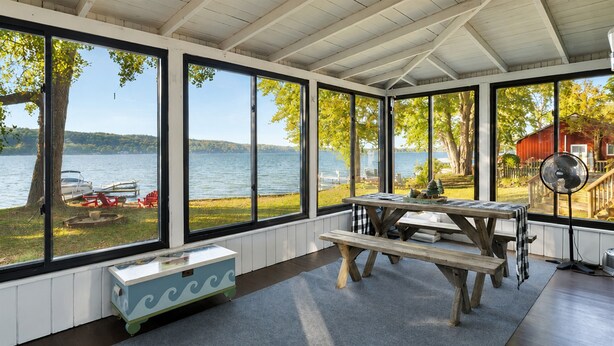 A covered patio with large windows overlooking a lake, a picnic table, and a fan. - Honeoye Cottage | 50' Lakefront | Dock | FLX (Honeoye)