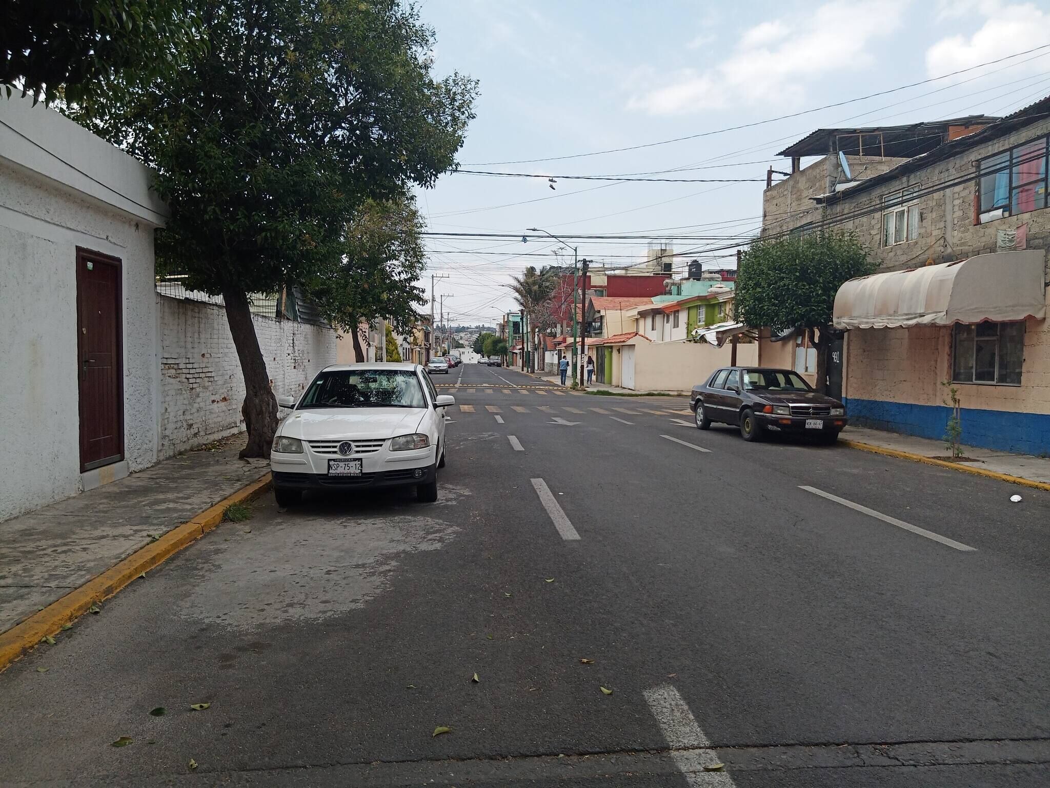 Una calle con edificios, un coche blanco estacionado al lado y un árbol.