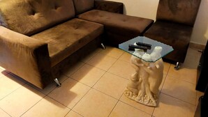 Una sala de estar con un sofá seccional marrón, una mesa de centro de vidrio con una escultura de árbol y una mesita con dos controles remotos.