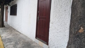 Un edificio blanco con una puerta marrón, una ventana con celosía metálica y un árbol al lado.