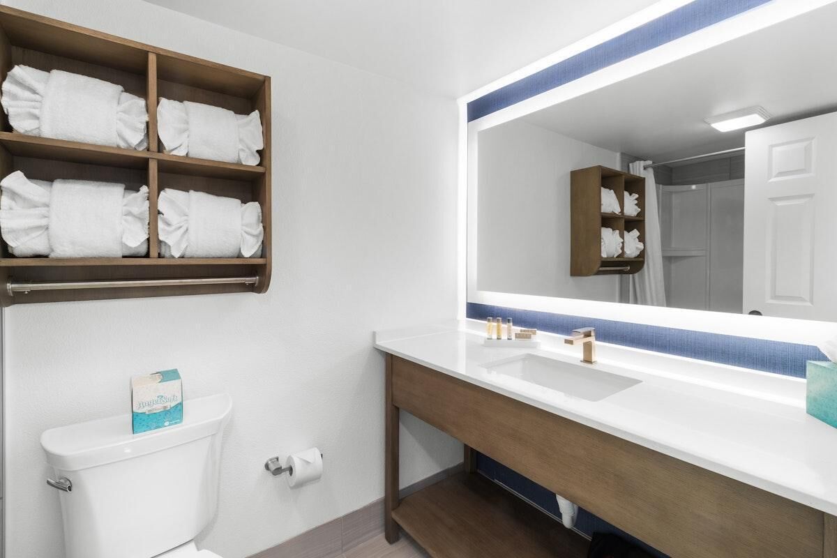 A bathroom with a white toilet, a wooden shelf with folded towels, a large mirror with a blue frame, and a white sink with a wooden vanity.
