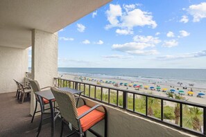A balcony with wicker chairs and a table, overlooking a beach with umbrellas and people.