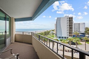 A balcony with a view of the beach and ocean, a chair, and a building with multiple floors in the background.
