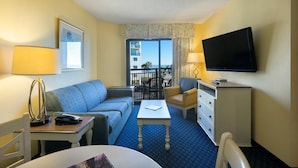 A hotel room with a blue carpet, a sofa, a chair, a TV, a desk, and a balcony with a view.