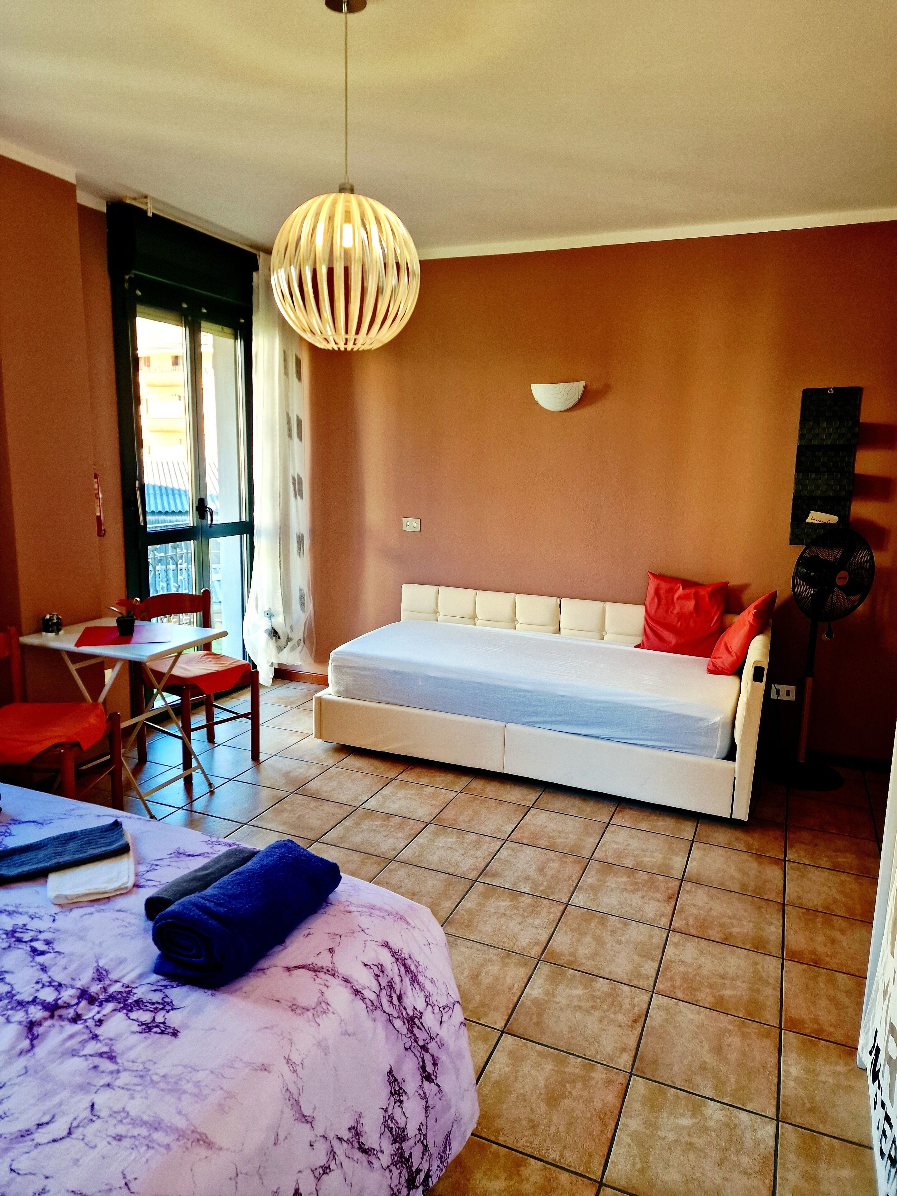 Ein Zimmer mit einem Bett, einem Esstisch, Stühlen, einer Pendelleuchte und einem Fenster.