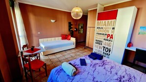 A hotel room with a bed, a sofa, a dining table, and a chair. There is a large wall with city names and a red suitcase on a shelf. - Bernina Station Suite: 30 Steps to the Red Train (Tirano)