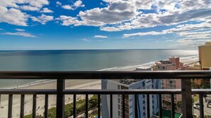 Udsigt over kysten fra en balkon med udsigt til strand og hav, med bygninger i baggrunden.