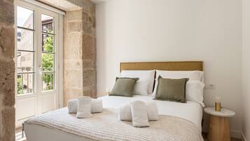 A bedroom with a large bed, a stone wall, and a window with a view of buildings.
