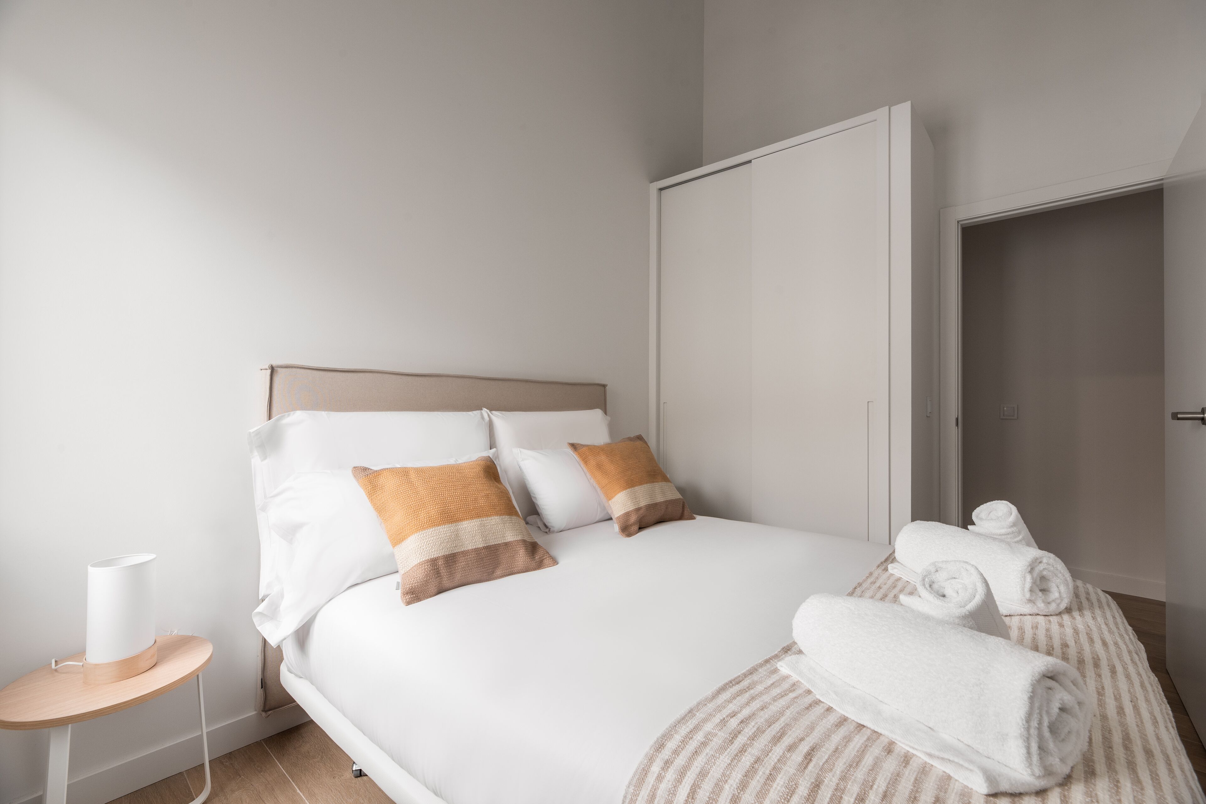 A neatly made bed with white and brown pillows, a small bedside table with a lamp, and rolled towels on a striped rug.
