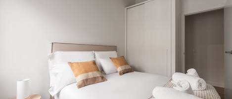 A neatly made bed with white and brown pillows, a small bedside table with a lamp, and rolled towels on a striped rug.