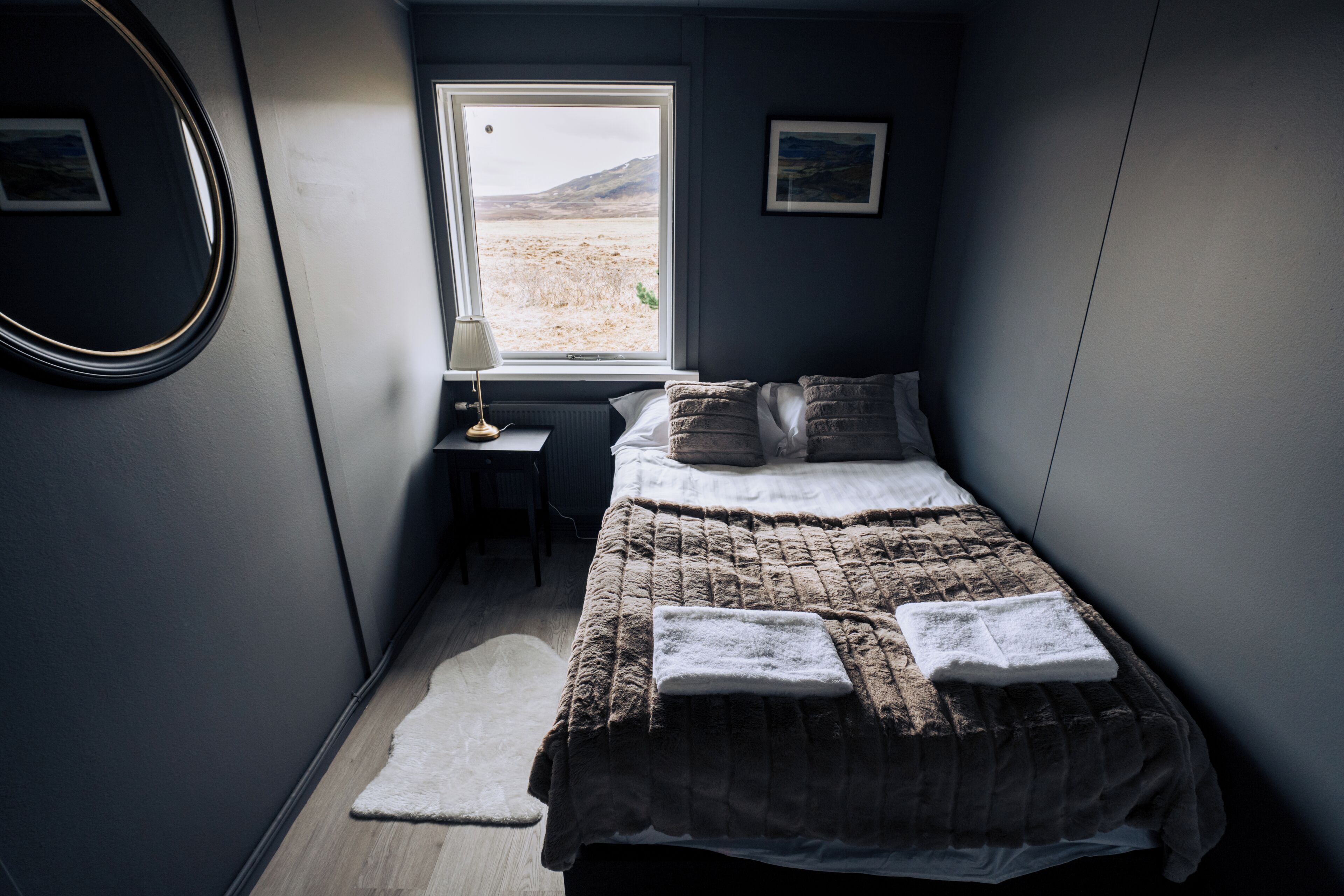 A bedroom with a bed, a small table, a lamp, and a window with a view of a desert landscape.