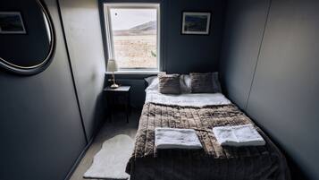 A bedroom with a bed, a small table, a lamp, and a window with a view of a desert landscape.
