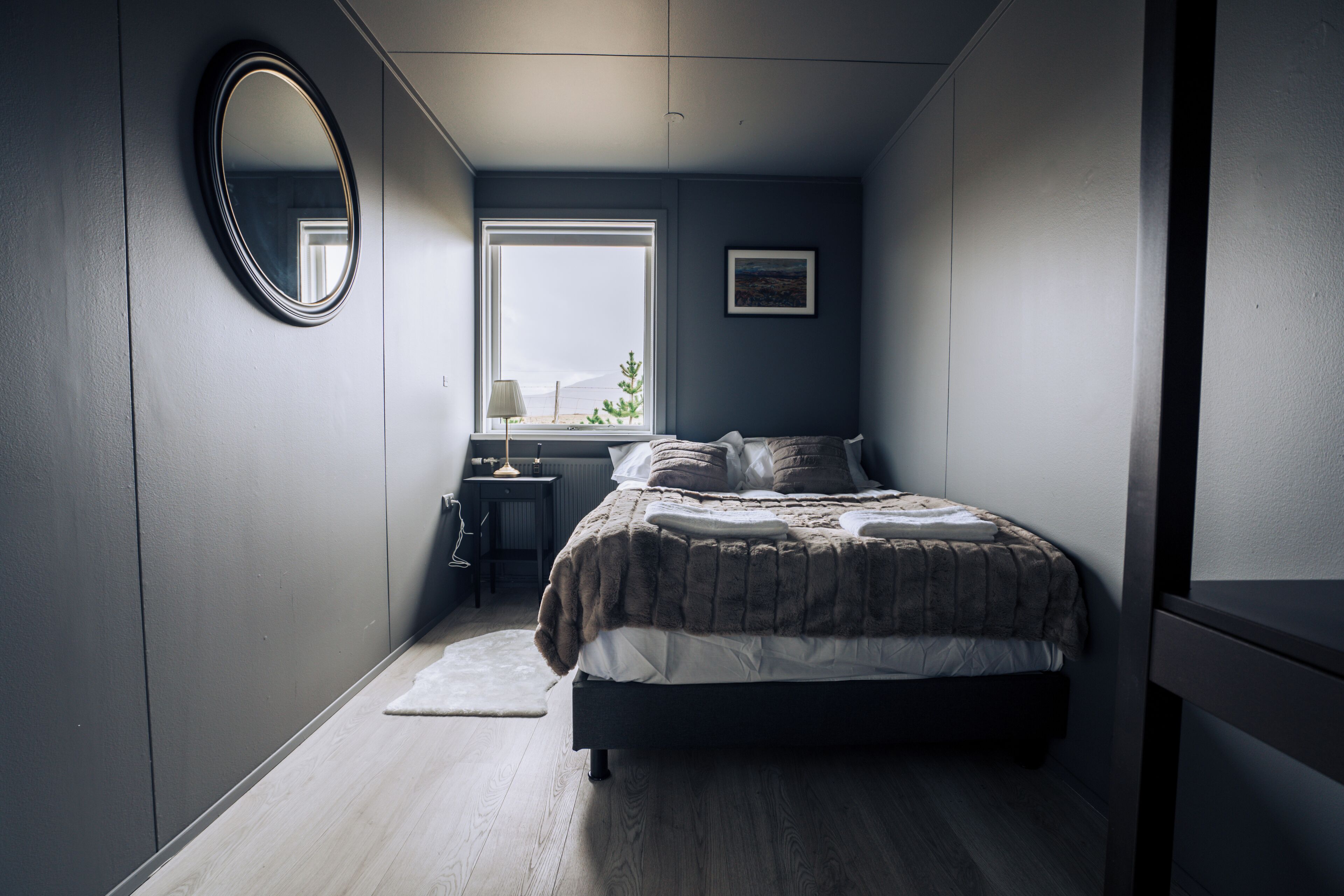 A bedroom with a bed, a bedside table, a lamp, a window, and a mirror.
