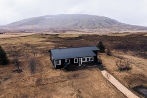 A black house with a sloped roof in a rural setting, surrounded by dry grassland and a mountain in the background. - Golden Circle Luxury Cottages (Selfoss)