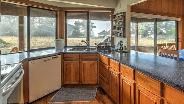 A kitchen with wooden cabinets, a built-in dishwasher, and a large window overlooking a yard. - Seabbatical | Cozy Coastal Getaway with EV Charger (The Sea Ranch)