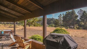 An outdoor deck with wooden chairs and a barbecue grill, overlooking a natural landscape with trees and a clear sky. - Seabbatical | Cozy Coastal Getaway with EV Charger (The Sea Ranch)