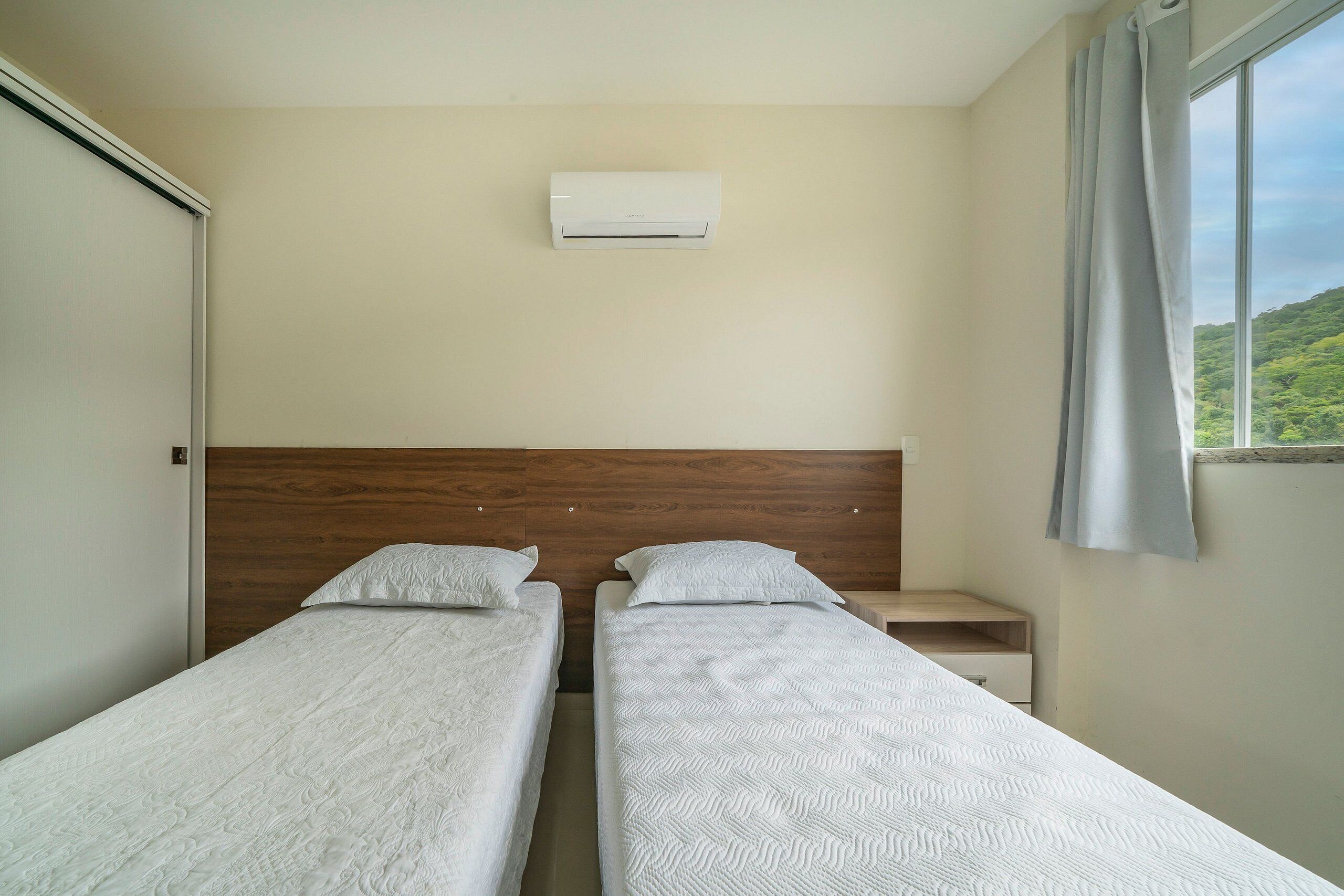 Een kamer met twee eenpersoonsbedden, een houten hoofdbord, een klein nachtkastje, een wandairconditioner en een raam met uitzicht op groen.