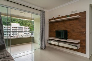 A modern living room with a glass door leading to a balcony, a flat-screen TV mounted on a wooden panel, and a minimalist design.