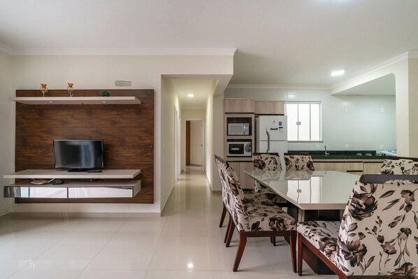 Una cocina moderna con electrodomésticos integrados, una zona de comedor con sillas de flores, y un televisor sobre una estantería de madera.