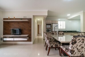 A modern kitchen with built-in appliances, a dining area with floral chairs, and a TV on a wooden shelf.