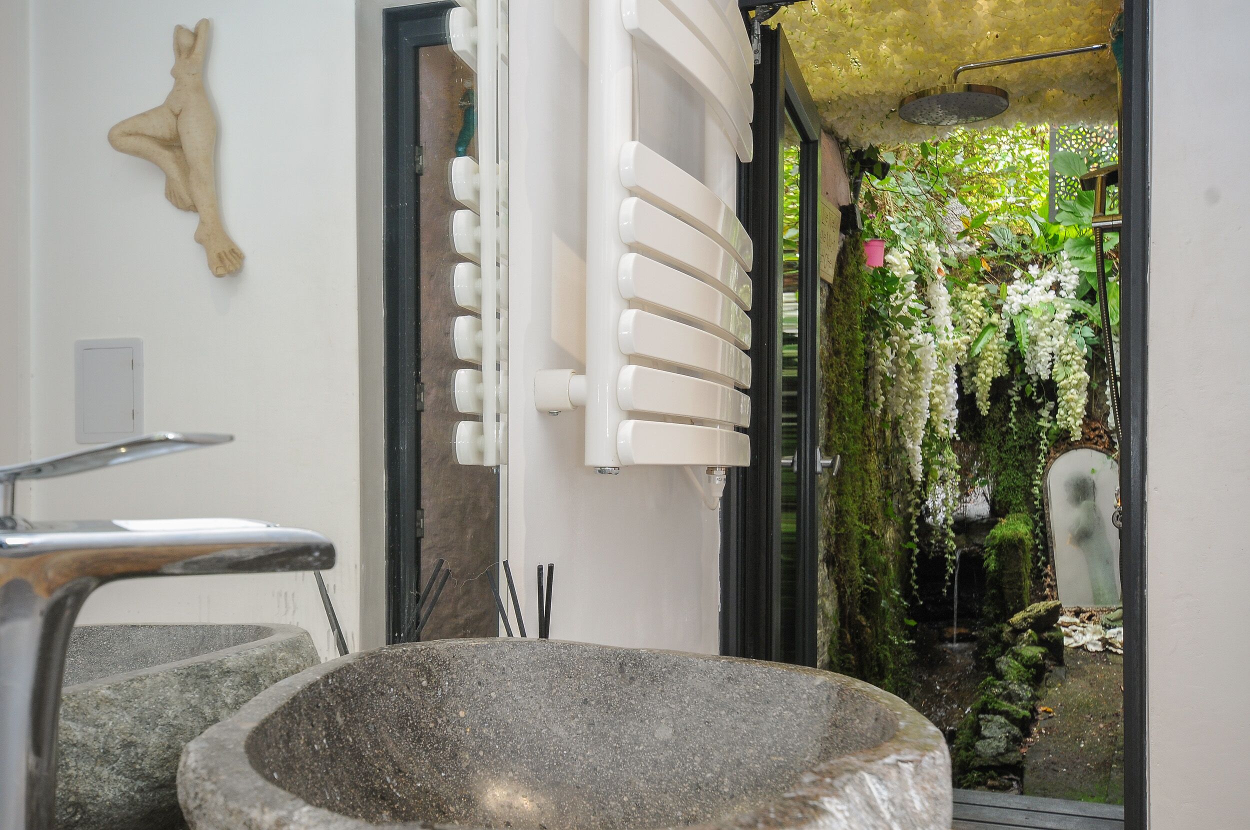 Un bagno con lavabo in pietra, termosifone bianco e vista su un giardino con fiori pendenti.
