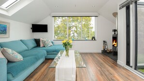 Een moderne woonkamer met een blauwgroene bank, een glazen salontafel, een open haard en een groot raam met uitzicht op bomen.