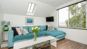 Een moderne woonkamer met een blauwgroene bank, een glazen salontafel, een groene vaas met gele bloemen, een schilderij en een aan de muur bevestigde televisie.