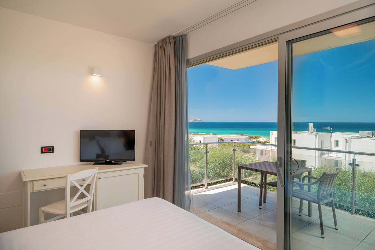 Une chambre d’hôtel comprenant un lit, un bureau avec une télévision, un balcon avec une table et des chaises, et une vue sur la mer.