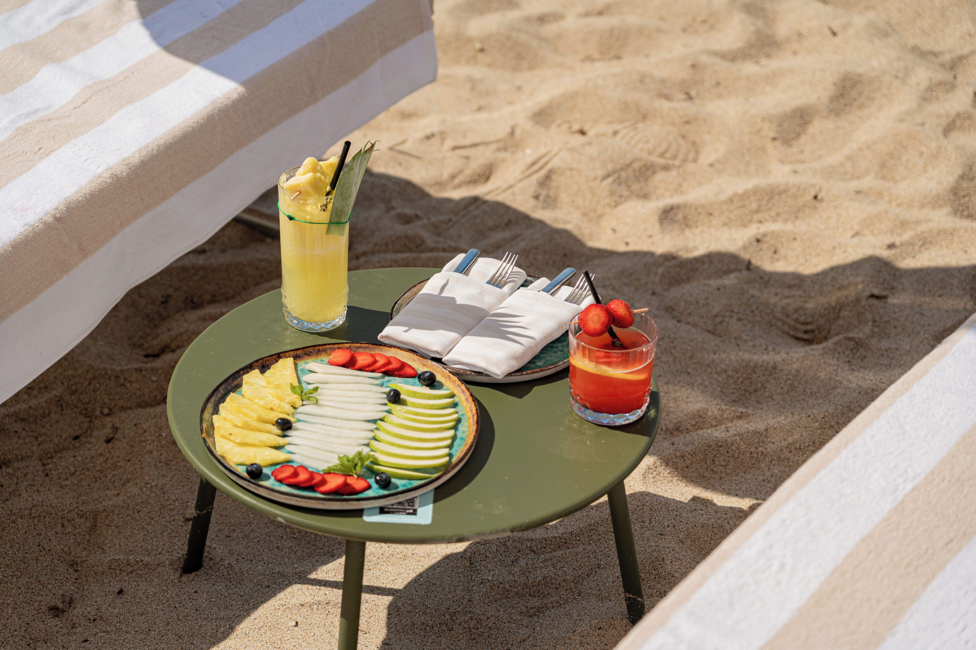 A table with a fruit platter, a glass of juice, and a drink on a beach setting.