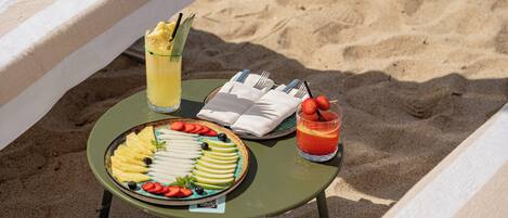 A table with a fruit platter, a glass of juice, and a drink on a beach setting.