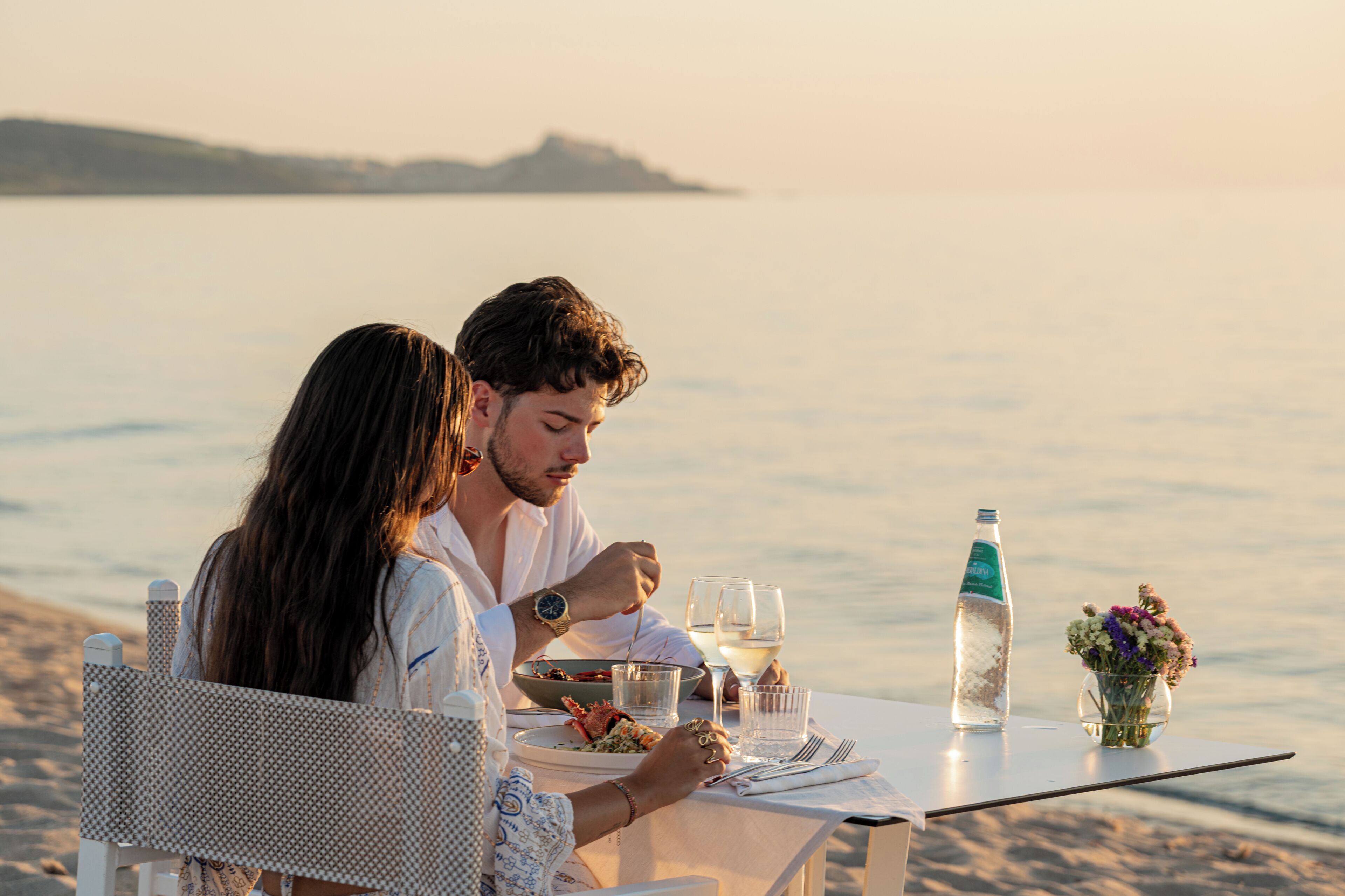 A couple dining at a table by the sea with a bottle of water and a vase of flowers.