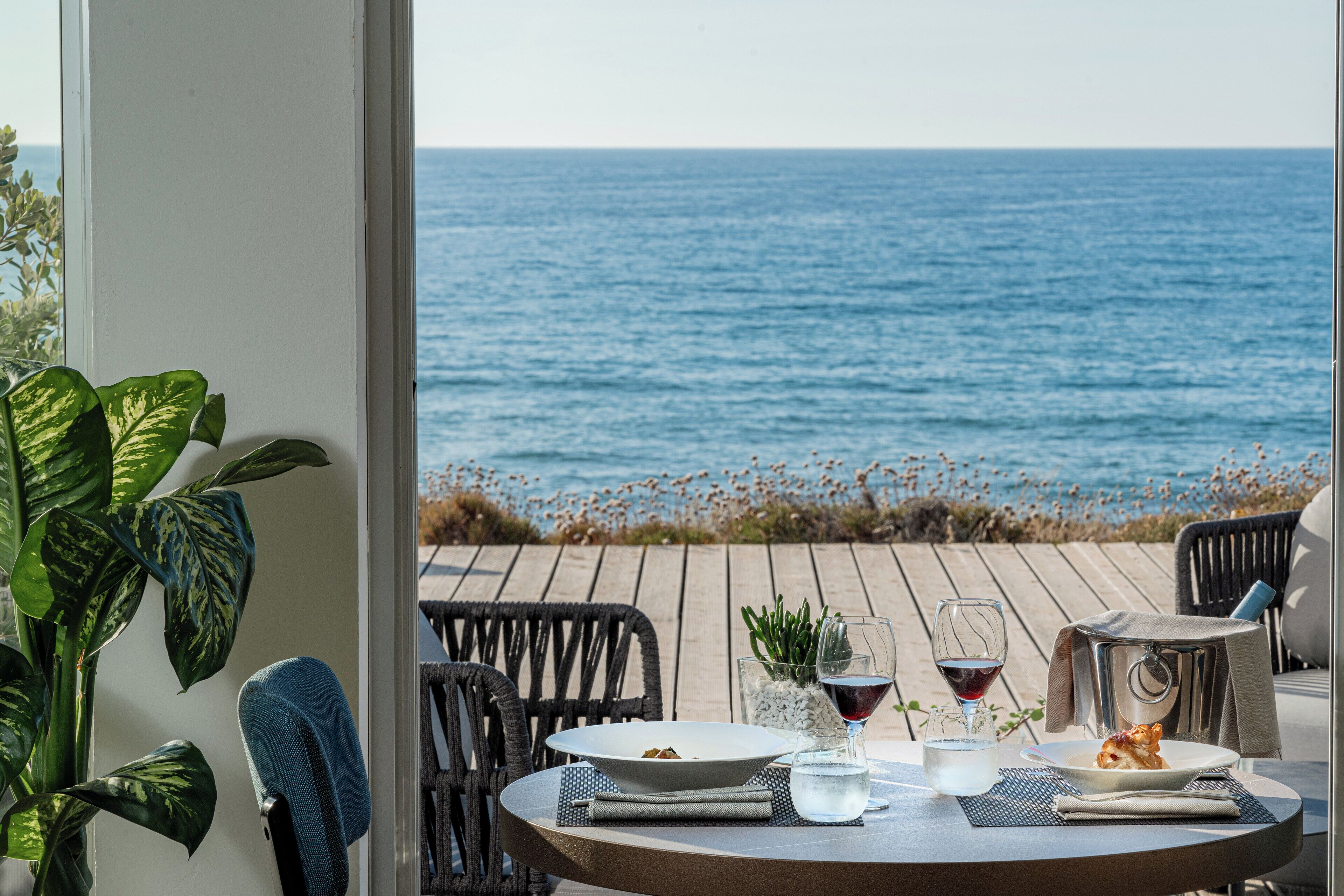 A dining table with a view of the ocean, a plant, and a glass of wine.