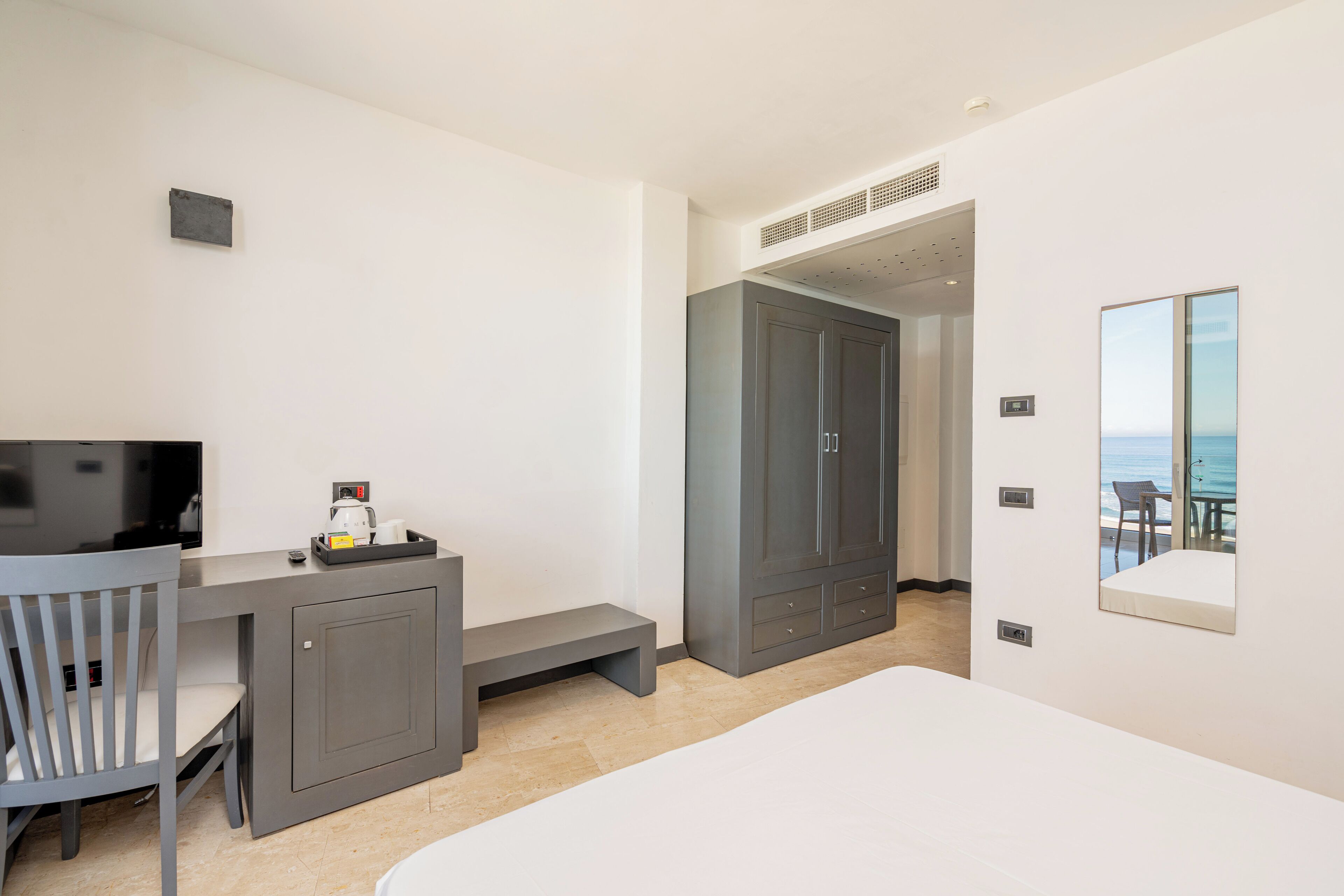 A modern hotel room with a bed, a desk with a chair, a wardrobe, and a view of the sea through a window.
