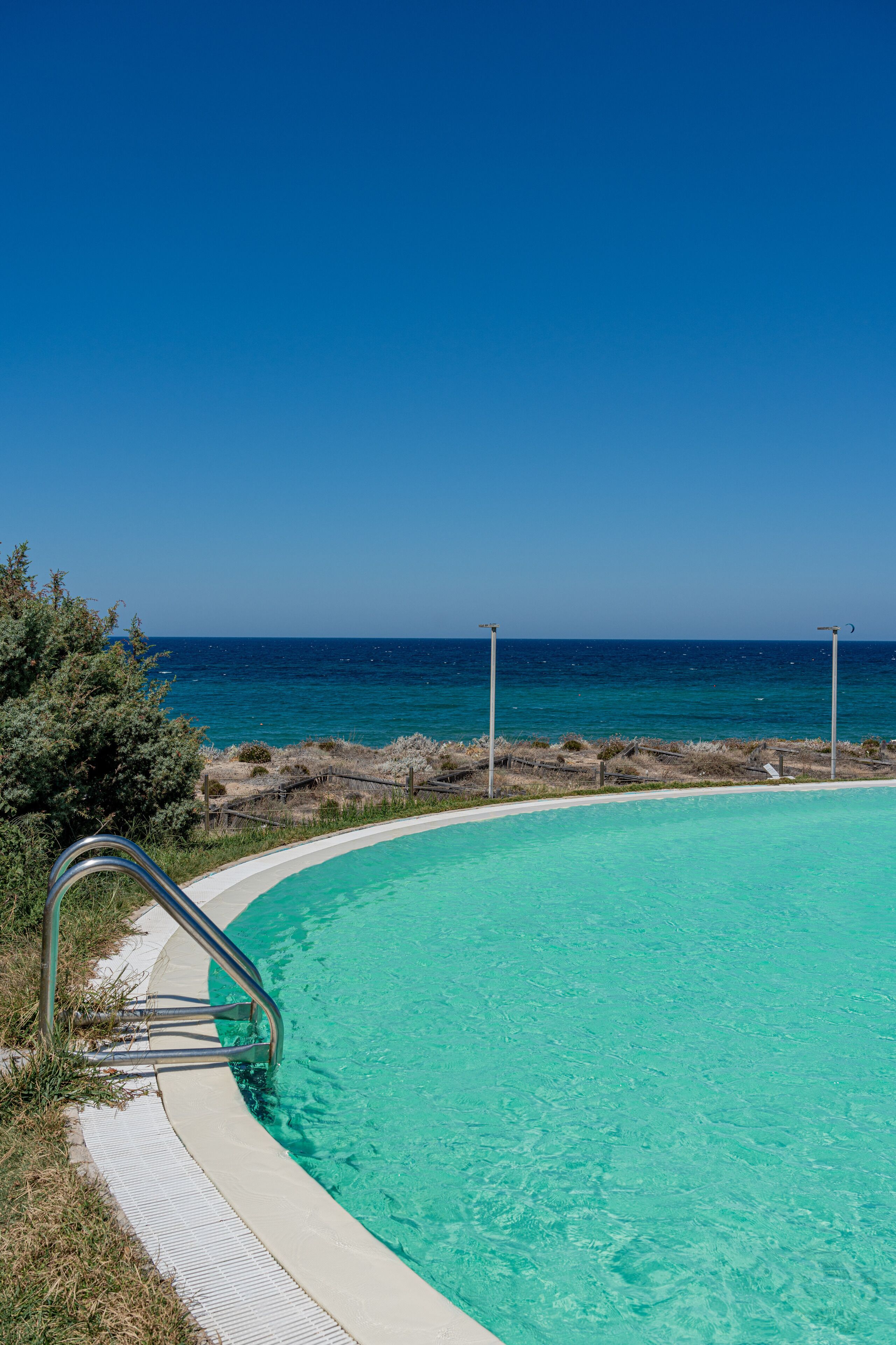 A clear blue pool with a metal ladder leading into it, surrounded by a white deck and overlooking a rocky coastline.