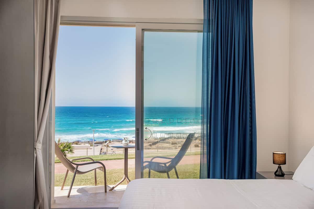 Une chambre d’hôtel avec une grande porte-fenêtre coulissante donnant sur la plage, un lit, une chaise et une table.