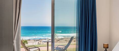 A hotel room with a large sliding glass door to the beach, a bed, a chair, and a table.