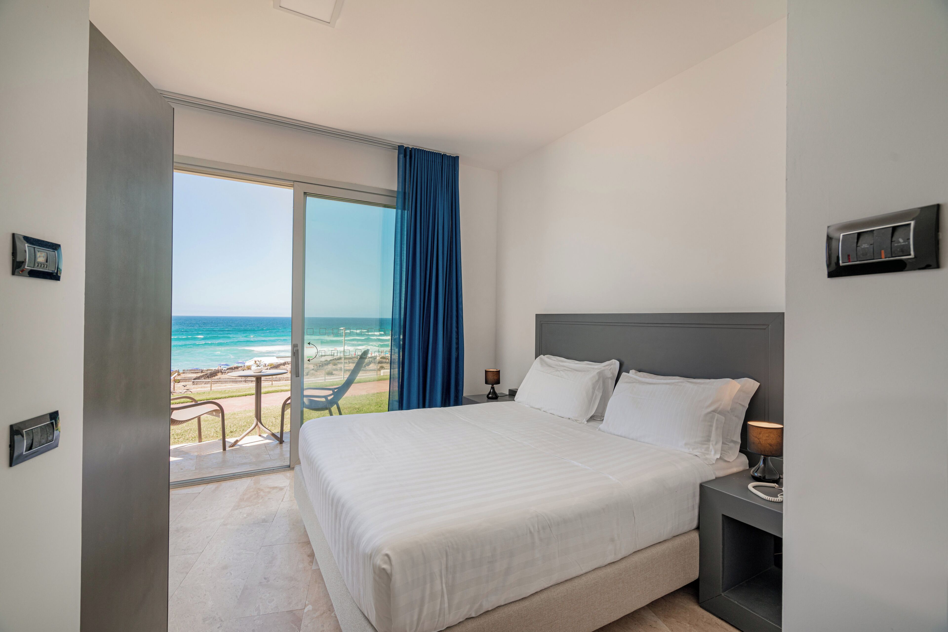 A hotel room with a large bed, a nightstand, a lamp, a view of the beach, and a sliding door.