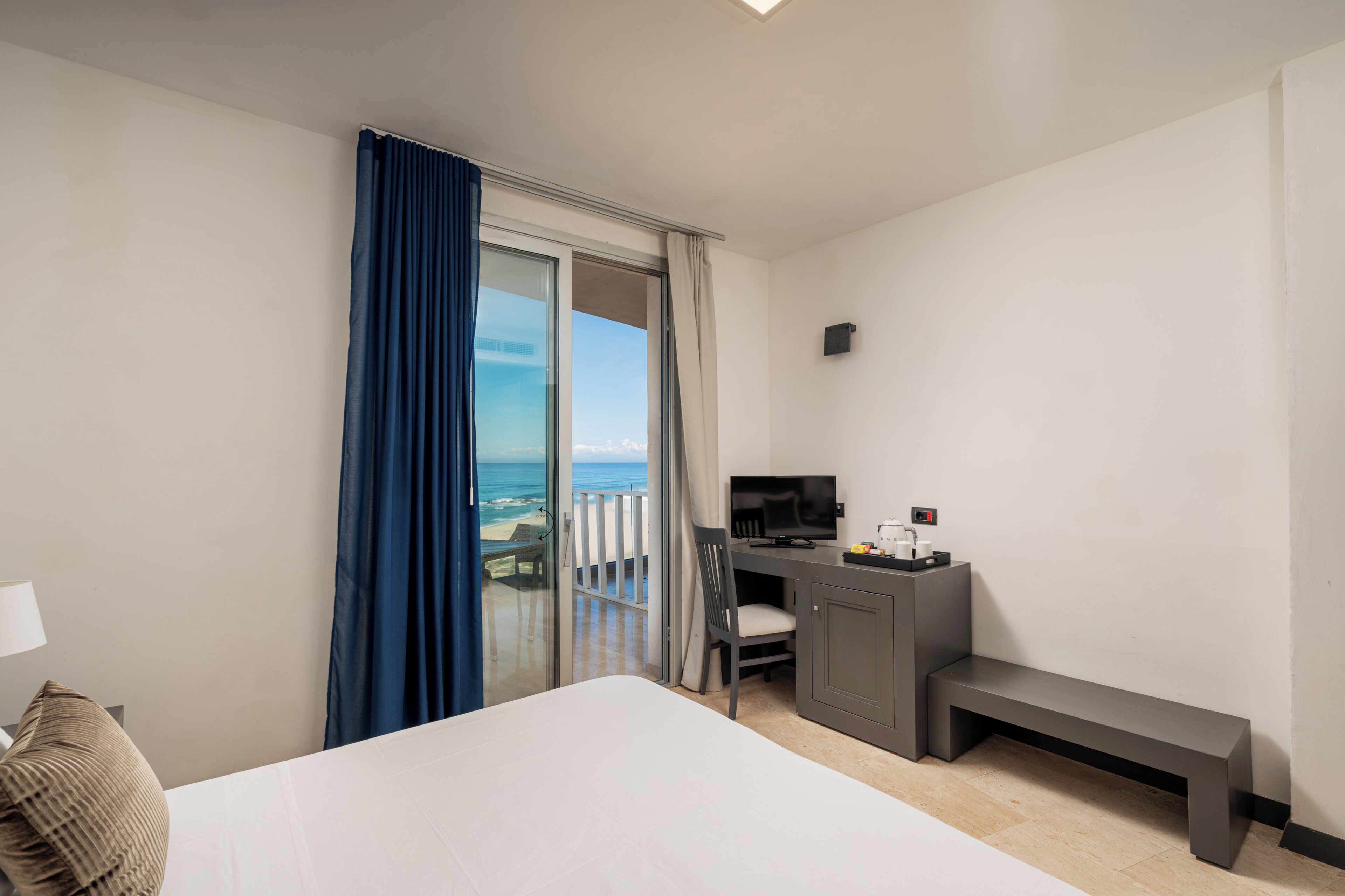 A hotel room with a bed, a desk with a chair, a TV, and a view of the ocean through a sliding door.