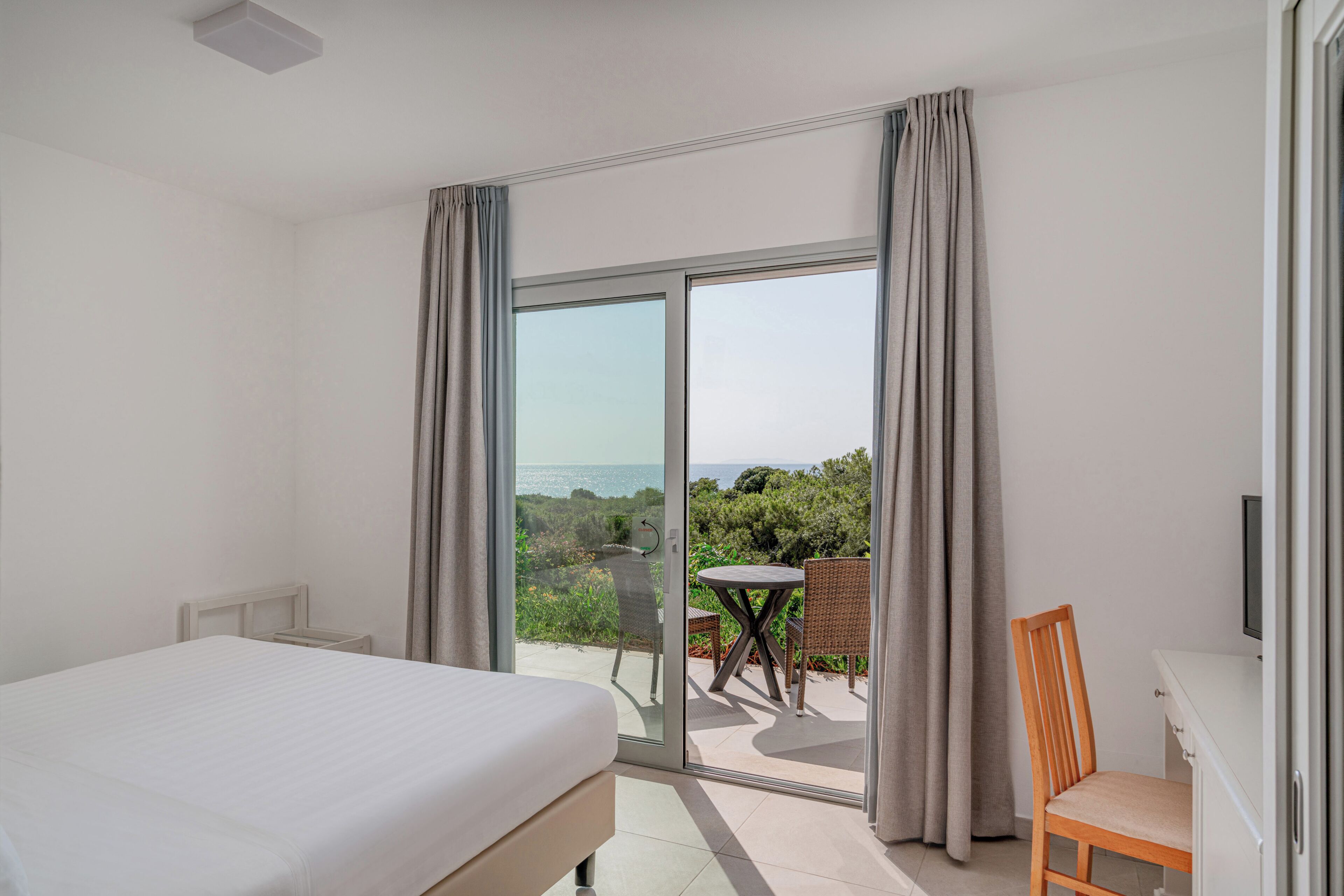A bedroom with a bed, a chair, a table, and a sliding glass door leading to an outdoor area with a view.