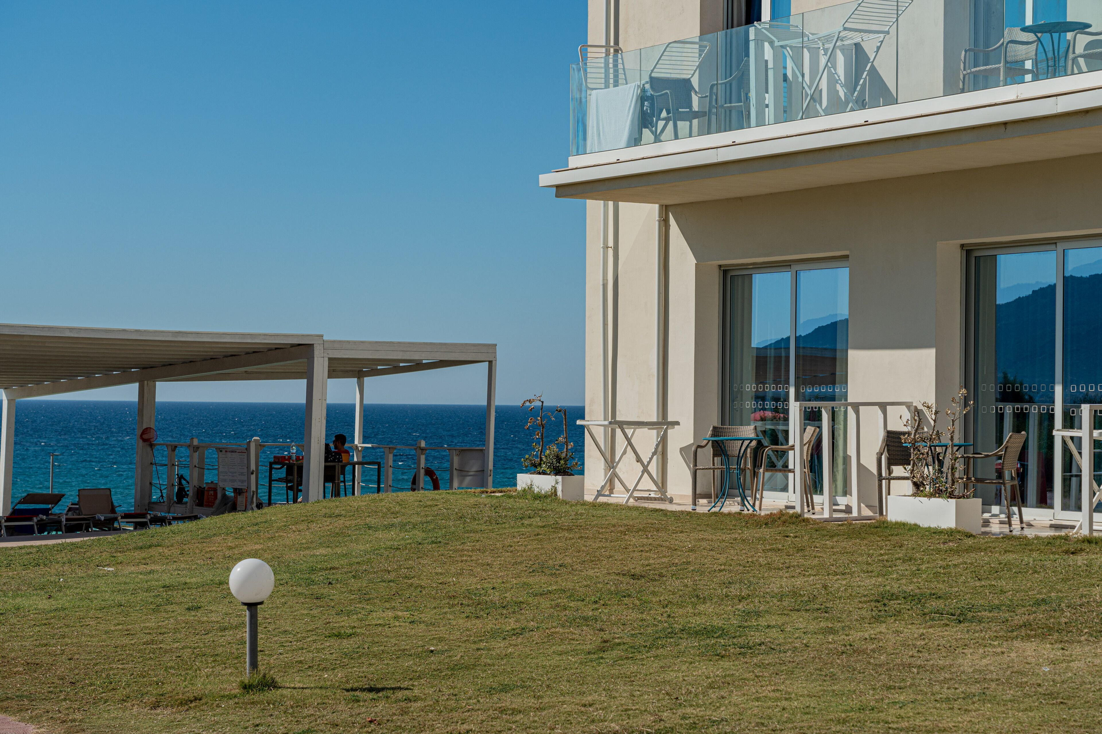 Ein modernes Gebäude mit Glasbalkon und Meerblick, Sitzgelegenheiten im Freien sowie einem überdachten Bereich mit Liegestühlen.