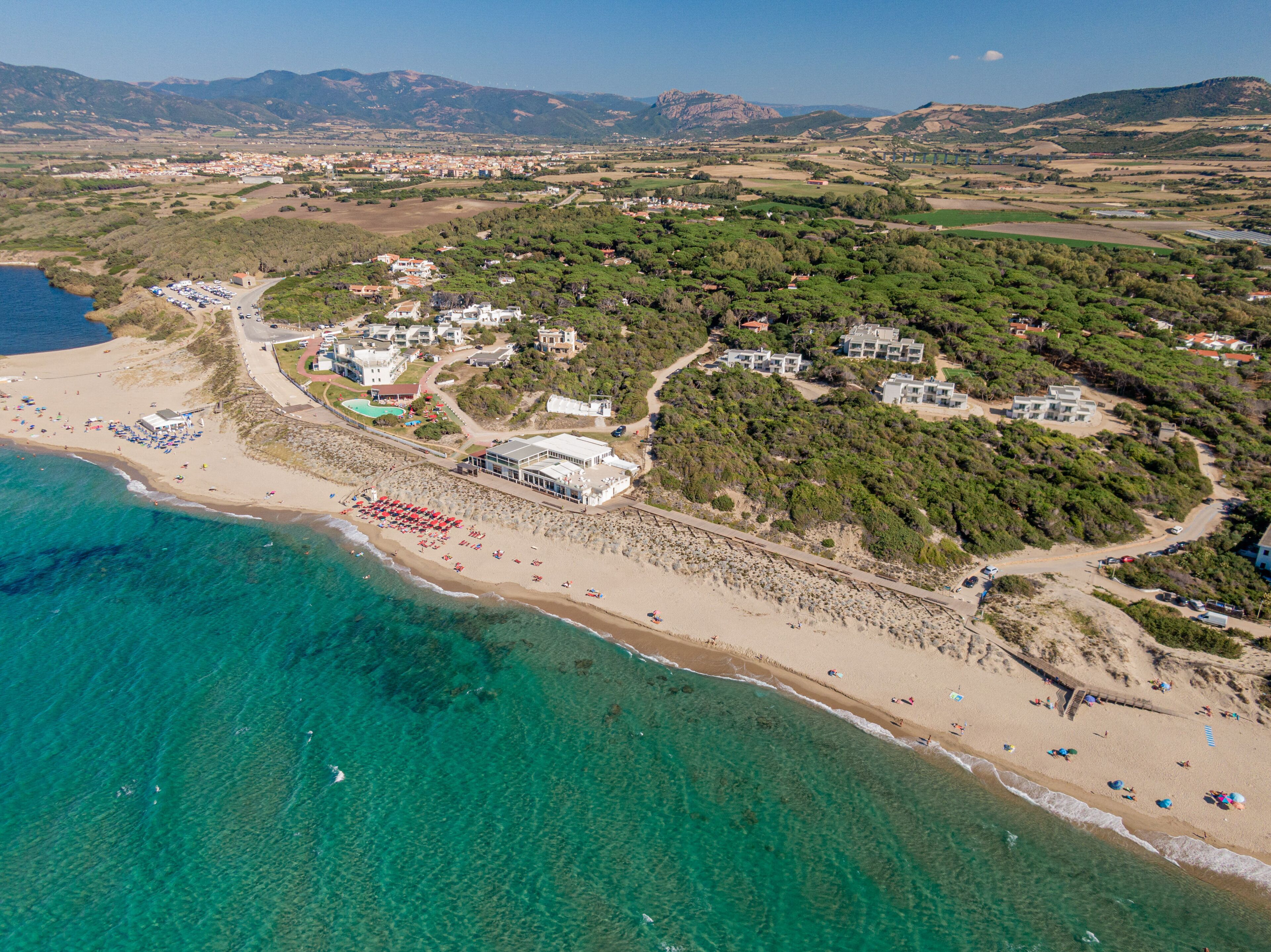 An aerial view of a coastal area with a beach, a small town, and a mountain range in the background.