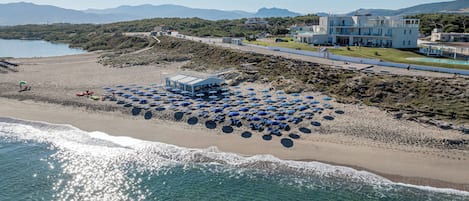 Une vue aérienne d’une plage, avec une rangée de parasols bleus et un immeuble doté de nombreux balcons.