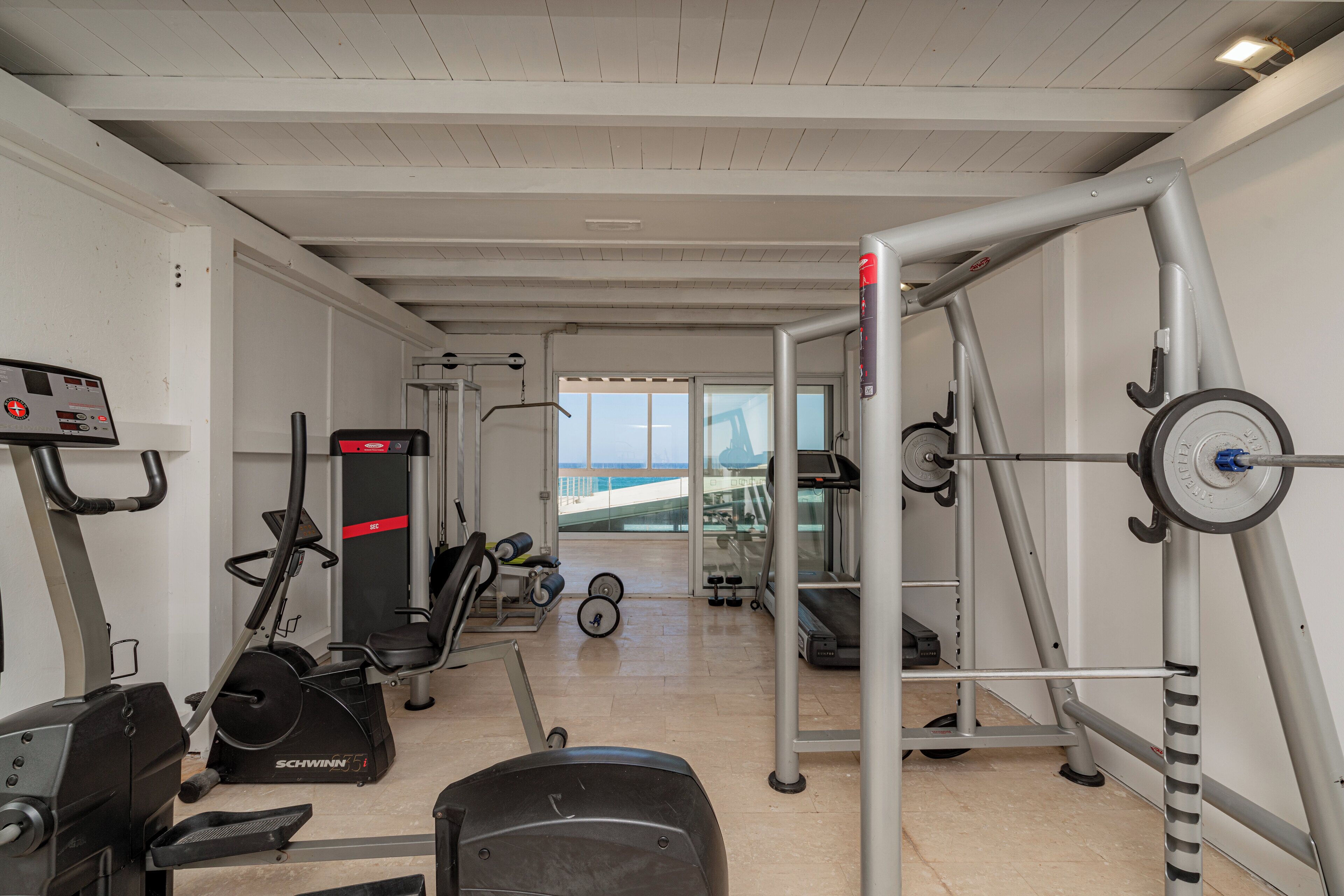 Une salle de sport équipée de machines de cardio, de matériel de musculation et offrant une vue sur la mer grâce à de grandes fenêtres.