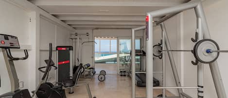 Une salle de sport équipée de machines de cardio, de matériel de musculation et offrant une vue sur la mer grâce à de grandes fenêtres.