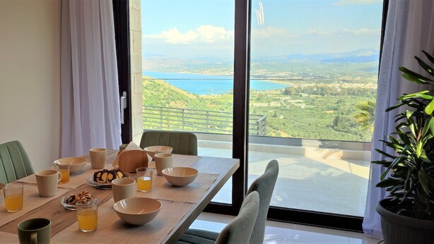 A dining table with bowls, glasses, and a plate of food, set against a scenic view of a lake and hills. - Villa ArGia with private pool & sea view (Apokoronas)