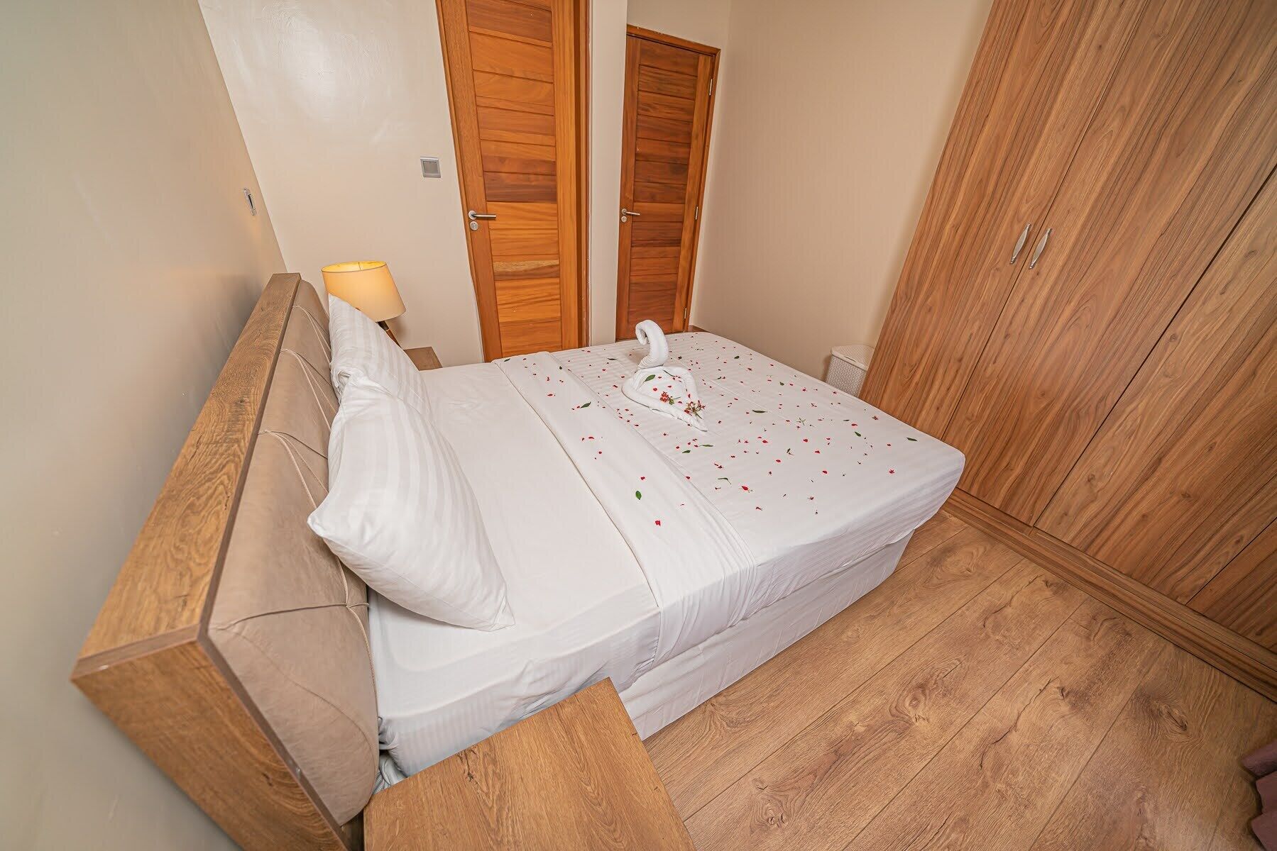 A neatly made bed with a swan-shaped pillow and a floral arrangement, wooden wardrobe, and a bedside lamp in a room with wooden flooring.