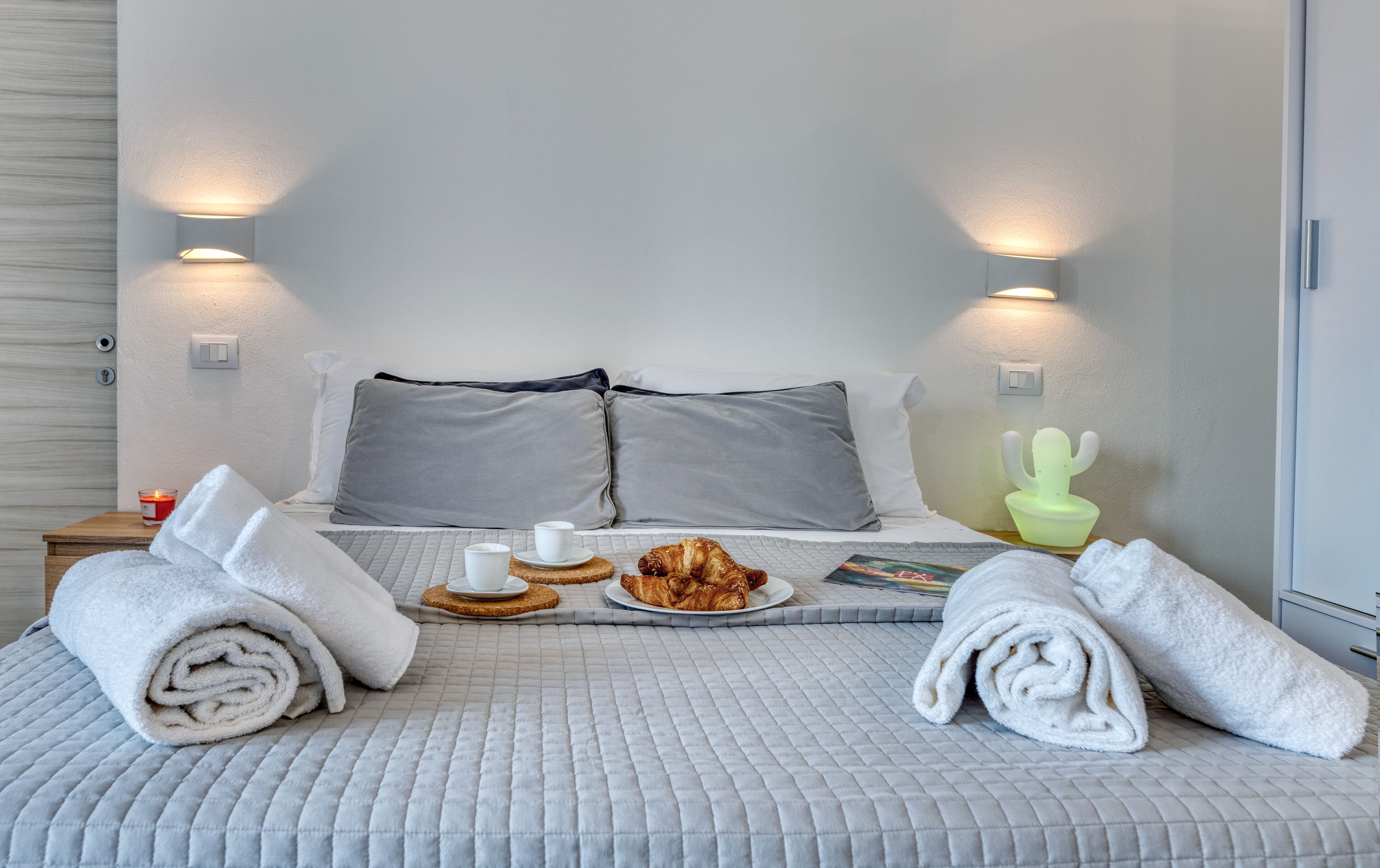A neatly made bed with pillows, a tray with croissants and cups, and a cactus-shaped nightlight.