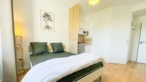 Een slaapkamer met een bed, nachtkastje, lamp, wanddecoratie, een keukenhoekje en een deur.