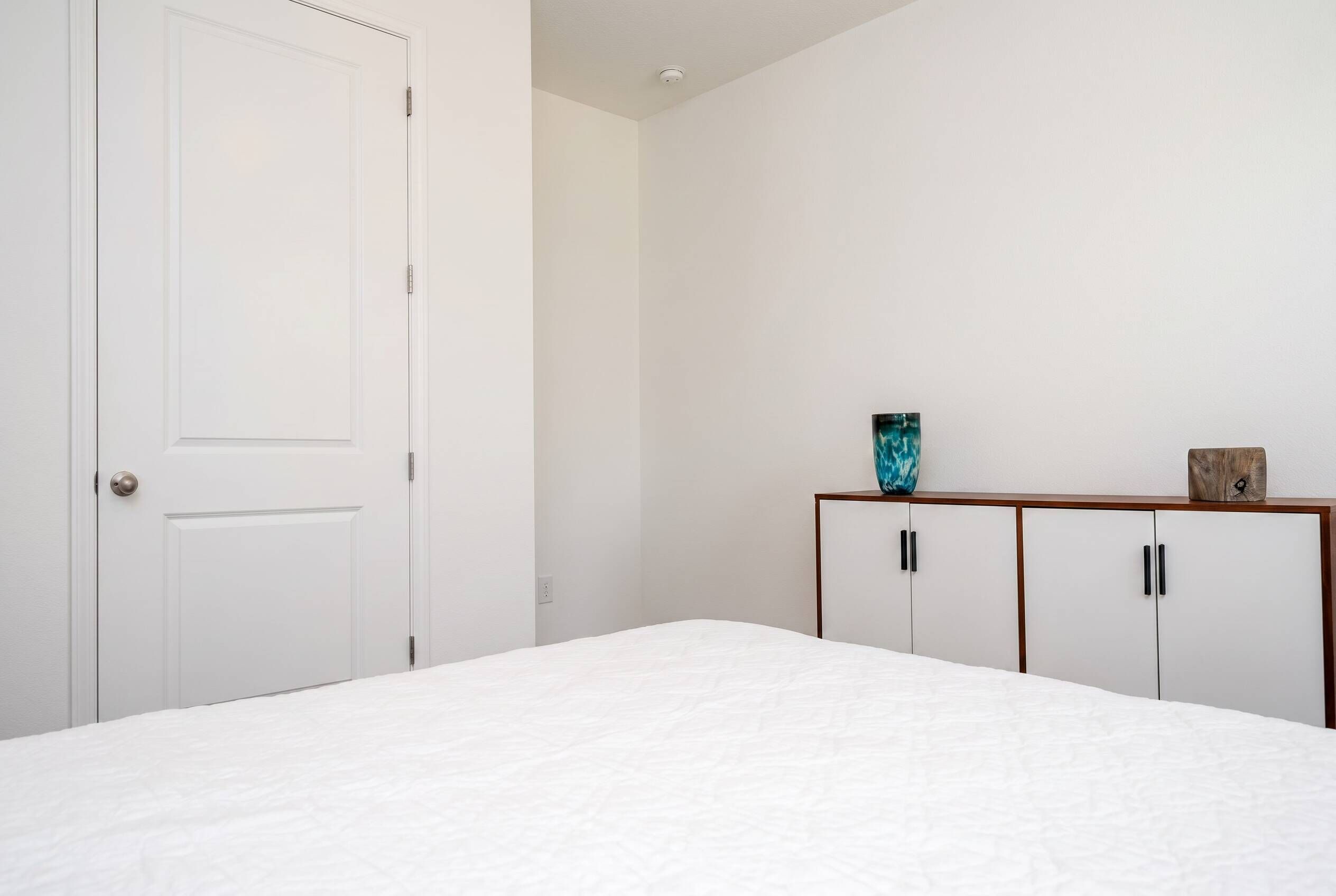 Een slaapkamer met een bed, een witte deur, een houten kast met zwarte handgrepen en een klein plankje met decoratieve objecten.